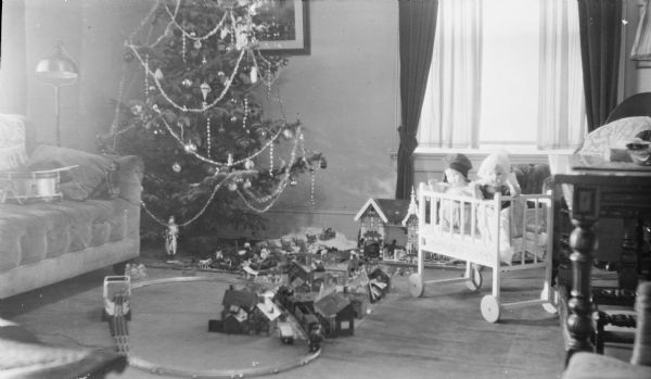 A Christmas tree stands in the corner of a living room furnished with a tufted sofa, left, and harp floor lamp. On the far right is a desk or writing table. In front of the tree is a toy train set and a village of small houses. There is a large doll house at the rear, partially obscured by a toy crib holding two large baby dolls. A Santa Claus figure is under the tree, which is decorated with ornaments, tinsel garland, and short lengths of glass bead garland.