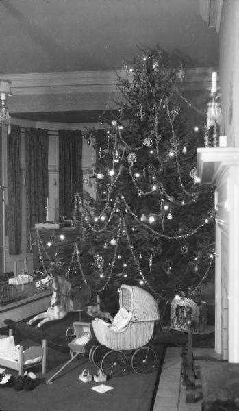 A tall Christmas tree with electric lights stands in the dining room of the Herbert P. Brumder residence at 2030 East Lafayette Place. The tree is decorated with glass and tinsel ornaments as well as tinsel garland. A hobby horse and doll baby buggy are in front of the tree. There is a die cut paper nativity scene on the floor at right.