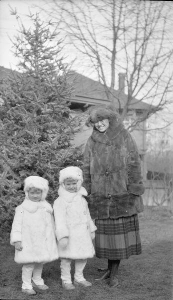 Margaret Bouer (Mrs. Herbert P.) Brumder posing outside the family house at 2030 East Lafayette Place with her two older children, Barbara, left, and Herbert Edmund. The children are dressed in matching outfits with white fur hats and coats, with white buttoned gaiters. Margaret is wearing a brimmed hat with feathers, a fur coat and pleated skirt. There is a large house in the background.