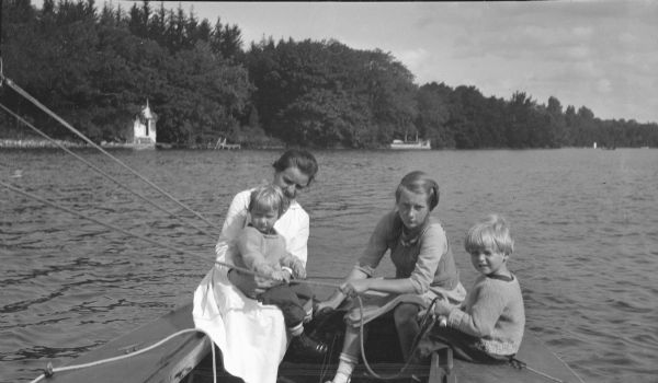 An unidentified young woman, left, is holding Barbara Brumder on her lap in the stern of a sailboat. A second young woman is holding a sheet (rope) with assistance from Herbert E. Brumder, far right. There is a boathouse on the shore at left, and the wharf at Villa Henrietta is at right, with two lamp posts.   