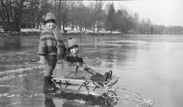 Two children, possibly Barbara, left, and Philip Brumder posing with a sled on the ice of Pine Lake. The children are dressed in similar outfits of stocking caps, sweaters, scarves, snowpants and galoshes.