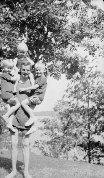 Herbert Paul Brumder posing with his three children near the shore of Pine Lake. He is standing holding Barbara, left, and Herbert Edmund, right, on his hips as Philip is sitting on his shoulders. They are all wearing swimming suits.
