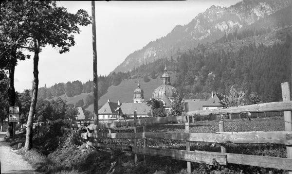 A view of the Benedictine monastery at Ettal, with the Bavarian Alps in the background. There is a fence in the foreground. Signs on a post at left identify the location, and advertise the availability of a "Telefon" at the Hotel Ludwig der Bayer.