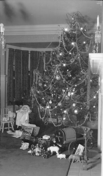 A tall Christmas tree with electric lights and tinsel garland stands in the dining room of the Herbert P. Brumder residence at 2030 East Lafayette Place. Toys have been placed on the floor in front of the tree and include two baby dolls, one in a cradle, a toy fire truck and a pedal car with windshield. There are also toy animals including a bear, pig and elephant.