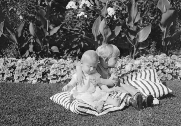 Herbert Edmund Brumder, right, posing with his arms around his sister Barbara, on the lawn at Villa Henrietta, the Brumder estate on Pine Lake. The children are sitting on striped pillows on the ground in front of a flower border with geraniums, cannas and garden phlox.
