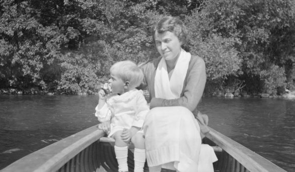 Margaret Bouer (Mrs. Herbert P.) Brumder sitting beside her son, Herbert E. Brumder in a canoe near the shore of Pine Lake. Herbert is eating an apple.