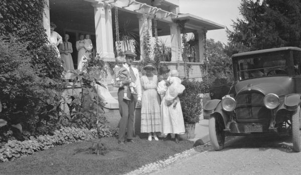 Herbert P. Brumder, wearing a cap and necktie, is holding his son Herbert E. Brumder on his hip while his wife, Margaret is holding baby Barbara. The woman between them is unidentified but may be Herbert's niece, Gertrude Merker Messinger. The group is standing near an automobile parked in front of the porch steps. Herbert's mother, Henriette Brumder, the widow of Milwaukee publisher and businessman George Brumder, is standing in the shade of the porch between two unidentified women, one of whom is holding an infant.