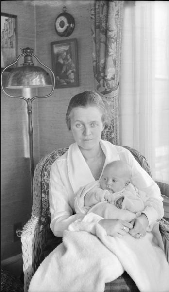 Gertrude Merker (Mrs. Clifford) Messinger sitting in an upholstered wicker chair next to a window, holding an infant, either her daughter Mary Baldwin Messinger, born 1918, or daughter Joanne Beecher Messinger, born 1920. There is a harp-style floor lamp with metal shade behind the chair.
