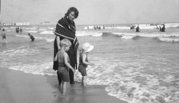 Margaret Bouer (Mrs. Herbert P.) Brumder is standing on a beach with her children Herbert E., left, and Barbara. The children are wearing matching swimsuits and Barbara has a bonnet. Margaret is also in swimming attire with a shawl around her shoulders. There are two billboards on a long pier in the background advertising Coca-Cola and Kelly Tires. A building with four short towers is standing at the end of the pier.