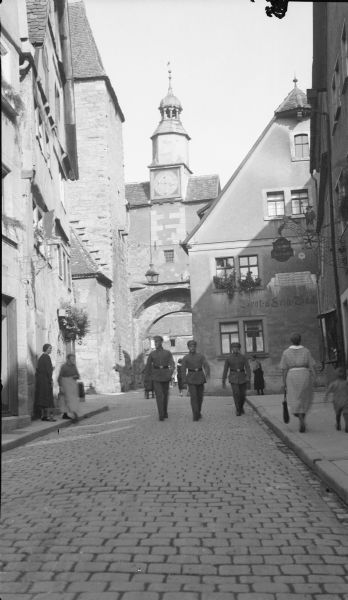 A view down the center of a street towards St. Mark's Tower in Rothenburg ob der Tauber, Germany. Three men in uniform are walking up the street. There are other pedestrians on the sidewalks. Ornate wrought iron signs are hanging from the facade of buildings on both sides of the street, and flowers are in bloom in several window boxes.  
