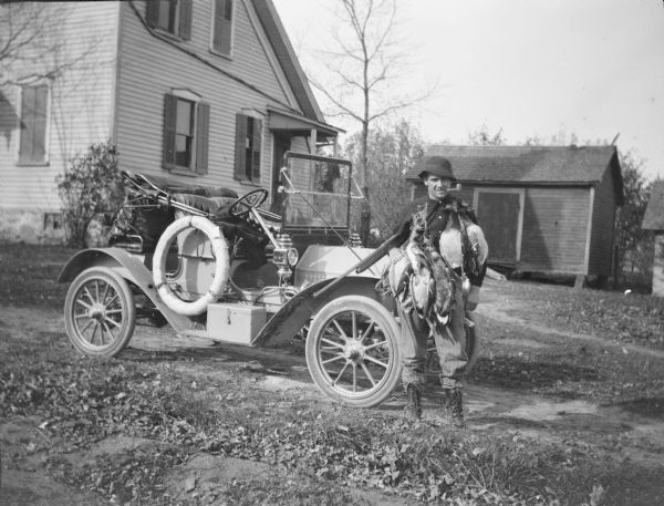 An unidentified man is standing beside an open roadster-style automobile. He is holding a shotgun and has a brace of ducks draped around his shoulders. The hunter is wearing a hat and has a corn cob pipe in his mouth. There is a saltbox style house on the left and a small shed in the background on the right. 