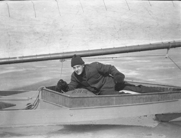 Herbert Paul Brumder reclining in the cockpit of an iceboat on Pine Lake. He is holding the main sheet in his right hand, and controlling the rudder with his left.