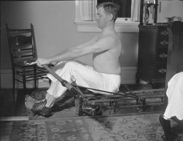 A profile view of Herbert Paul Brumder sitting shirtless on a rowing machine. There is an oriental-style carpet on the floor.