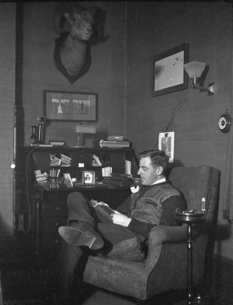Herbert Paul Brumder sitting and reading in an upholstered chair in front of a desk. He is smoking a pipe and there is a small smoking stand to the right of the chair. A neck mount of a bighorn sheep is hanging high on the wall above the desk. Below the mount is a double frame containing photographs of his mother, Henriette, and father, George. A telephone and desk lamp are sitting atop the desk.