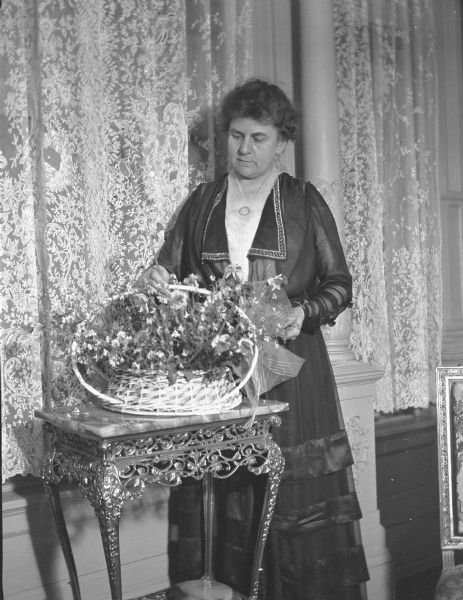Ida Brumder Merker is standing and admiring spring flowers arranged in a basket with a large ribbon bow. She is wearing a fancy dress and is holding a flower in her right hand. The basket is resting on an ornate marble topped table with a metal base. There are lace curtains and a column behind her.