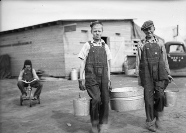 One of a group of images under the headline: "Matanuska Valley Colonists Wait for Mail, Carry Water, Haul Logs." Original caption reads: "You can't get water in Palmer by turning a faucet. These two boys are shown carrying water back to their tent in pails. Apparently having their picture taken made the task more enjoyable."