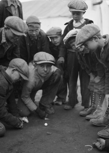 Group of boys playing marble game outdoors. One image under the headline: "The Kids Love Alaska; There's So Much to Do." Original caption reads: "Marbles supply one of the main diversions inthe headquarter camp. Here are some of the boys as they 'knuckled down' just as if they were back in the States."