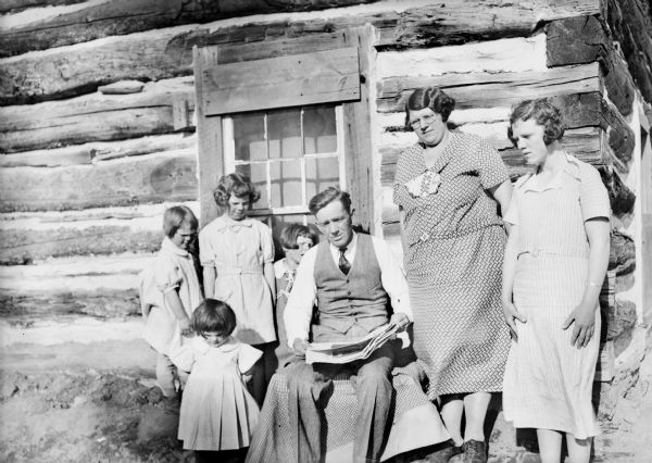 Family sitting outdoors. Original caption reads: "These are members of the first Wisconsin families named to go to Alaska in connection with the government's colonization of the Matanuska valley. A typical family is that of Henry James Roughan of Monico, Oneida county. He is shown reading a story of the venture to his wife and five daughters, Dona Mae, Carol, Beatrice, Bernice and Edith."