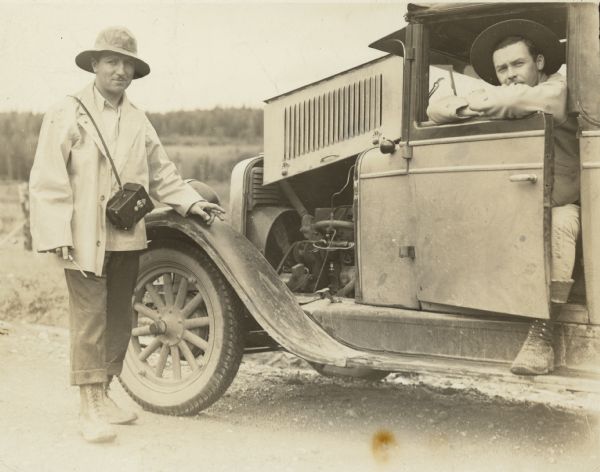 Arville Schaleben standing next to an automobile with a camera over his shoulder, a screwdriver in his right hand, and a cigarette in his left hand. A man is sitting in the driver's seat of the car leaning on the partially open door. The hood of the car is folded up over the engine. Alaska Colony. Caption on back of print reads: "Arv. Schalaben and friend in Matanuska Valley Alaska, 1935."