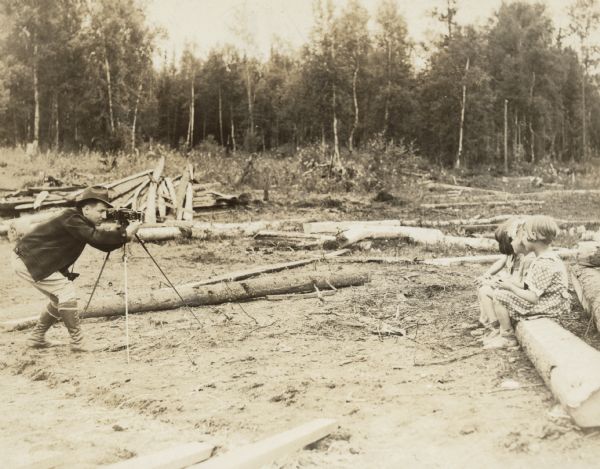 Arville Schaleben taking a photograph of three children sitting on a log with a camera on a tripod in the Alaska Colony. Caption on back of print reads: "Arv. Schalaben at Matanuska Valley in Alaska — 1935."