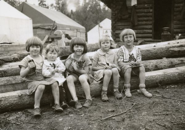 Group portrait of children sitting on a stack of logs. One of the children is holding a cat. Taken at Colony, Camp Number 5. Cabins are in the background. Caption from A.S. notes with negative: "Five little girls with nothing to do but look pleasant — Donna Mae Roughan, Shirley Herried, Berniece Roughan, Patricia Johnson, and Joyce Herried. All are from Wisconsin and all now live at Camp 5." 