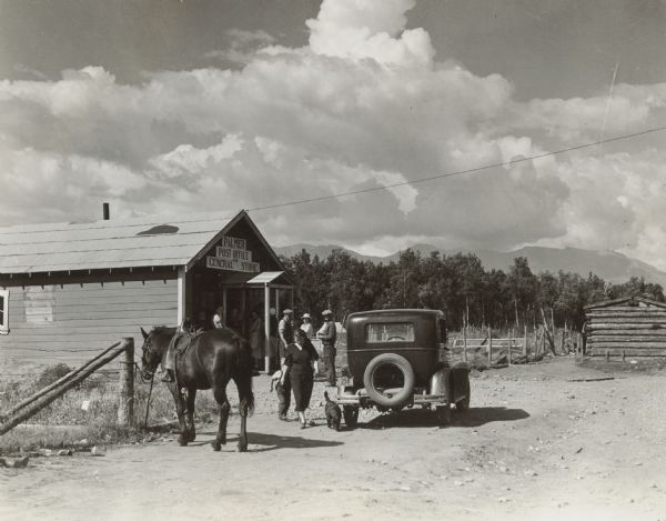 People are gathered at the entrance to the Post Office and General Store. There is a horse tethered to a wooden fence post, and an automobile is parked nearby. There are mountains in the background.