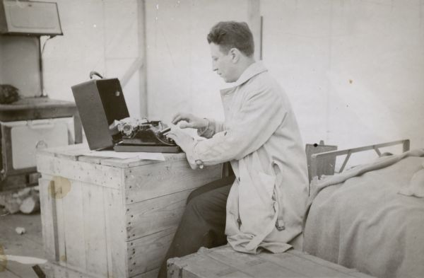 Arville Schaleben using a typewriter in a tent. Caption with negative reads: "One-second exposure of myself inside out [sic] sleeping quarters and office."