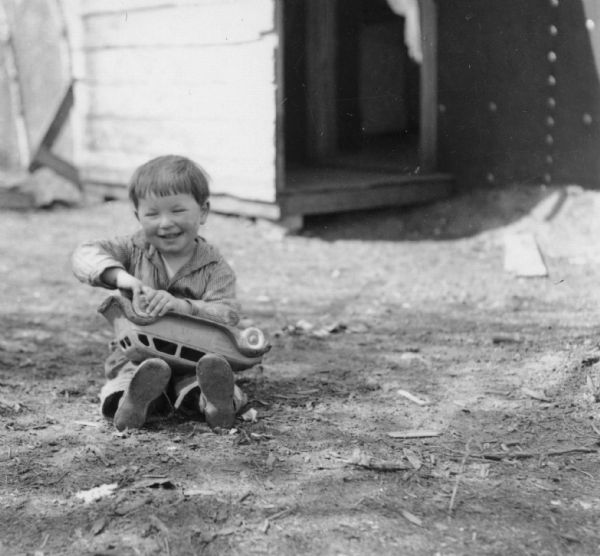 A boy is sitting on the ground outdoors, holding a toy car in his lap. Original caption reads: "Complaints that one doctor is not enough to care for the medical needs of the colonists at Palmer, Alaska, are being made of Martin Soyk, formerly of Minocqua, Wis., whose son Jimmy (above) is suffering from two diseases, chicken-pox and scarlet fever. Jimmy just recovered from a case of measles." Handwritten on back of print: "It's fun to be a kid. 'Wow, my own car!"