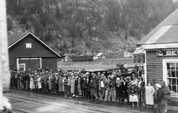 Group of people on dock to welcome the pioneers. A woman standing on the dock on the right appears to be holding a camera at her waist. One of a group of images under the headline: "First Pictures of Wisconsi's Alaskan Pioneers at Sea and Their Arrival at Seward." Original caption reads: "Here is the reception committee for the Wisconsin contingent of the pioneers. The picture shows the crowd on the pier at Seward waiting for the St. Mihiel to dock."