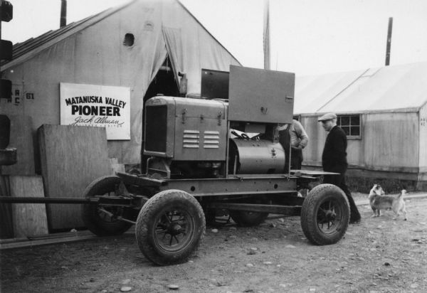 A vehicle or tractor on a trailer in front of a tent with a sign for "Matanuska Valley Pioneer, Jack Allman, Publisher • Editor." Two men and a dog are standing behind the trailer.