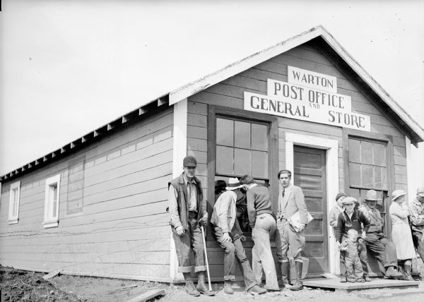 One of a group of images under the headline: "Matanuska Valley Colonists Wait for Mail, Carry Water, Haul Logs." Original caption reads: "Colonists lounge outside the general store and post office which they call Warton waiting for the postmaster to open the doors after separating the weekly mail. Some can't refrain from peeking in to see if they received a letter."