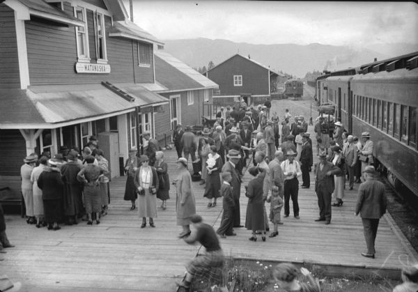 Elevated view of people on the platform at the railroad station. Caption with negative reads: "Alaska's tourist season at height — McKinley park route tourists stop for a few minutes at Matanuska village. Many of them catch a train there to visit valley colony."