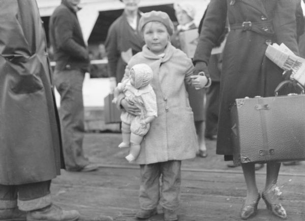 A young girl, Virginia La Rose, age 5, wearing a coat and hat and holding a doll under her right arm, is ready to disembark. She is the daughter of Henry and Clystia La Rose, originally from Price County, Wisconsin. A woman (face out of frame) standing next to her on the right, is carrying a newspaper and a piece of luggage, and is holding the young girls' left hand. Other people are standing in the background.