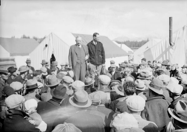 View over heads of crowd towards two men standing above the the crowd. There are tents behind them, and mountains in the distance. Caption could be: "Same as No. 7 with Don Irwin, in charge of project, shown at left. With Don is Col. O.F. Ohlson, general manager Alaska railroad, being introduced to settlers." Caption on print reads: "Col. Otto Ohlson (left) and Colony Manager Don Irwin explain how the colony men will draw lots for their 40-acre Matanuska Valley tracts."