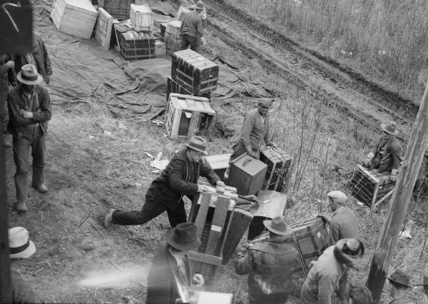 Elevated view of men working with the freight thrown off along the railroad right of way. Trunks and crates and barrels are arranged on a tarp in the background.