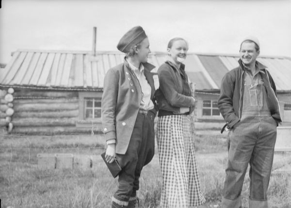 Marceline Venne, Betty and Fritz Hermon standing outdoors in front of a wire fence. There is a log building in the background.