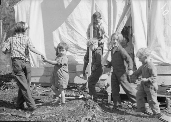 A group of young colonists in front of a tent. A person is standing just inside the partially open doorway of the tent. Caption on print reads: "Kids loved the Valley right from the start, with older ones readily helping the younger."