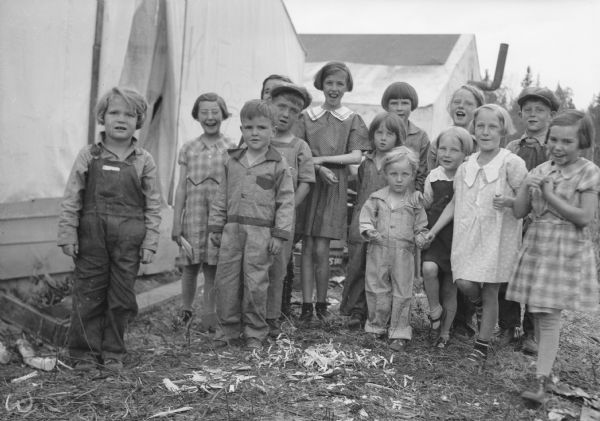A group of children are standing together near a tent. A young girl wearing overalls is standing on the left. Caption with negative reads: "'Surprise.' Children yell for fifth birthday of Evelyn Foster, from Stephenson, Mich. Evelyn at extreme left." Caption with print reads: "Who today remembers who in this wonderful cluster of pioneers 50 years ago?"