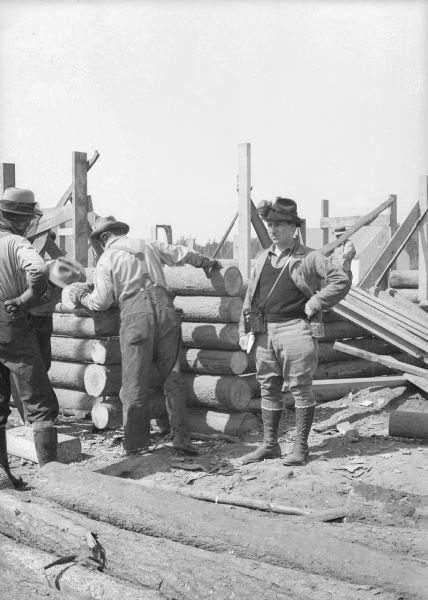 Arville Schaleben, with a camera case strung over his shoulders and a mosquito net on his hat is standing near workers who are constructing a log building. This may be the Construction Division office. Caption on print reads: "July 1935. Correspond[sic] Arville Schaleben 'supervises' making the first jail."