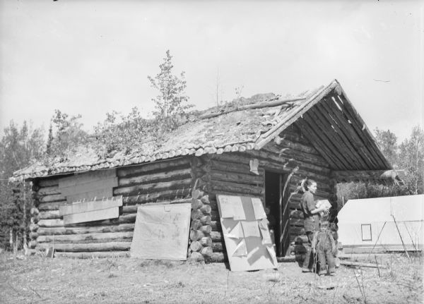 A woman and child are in front of a log cabin. There is a tent in the background on the right. Plants are growing on the roof of the cabin, and boards are covering up what may be a window on the left side. Caption with negative reads: "An abandoned log cabin is now commissary for camp 4. Mrs. Harry Campbell, [Abrams?], Wis., is leaving with groceries."