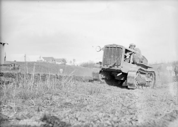 A man is driving a caterpillar tractor. A group of men are standing behind him on the right. On the left is part of a building and an agricultural implement, and in the distance is a person standing on a hill. A dwelling or farm buildings are on the hill in the distance. Caption with negative reads: "Pulling off topsoil with caterpillar tractor to get at gravel for gravelling roads."