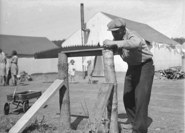 A man is standing and sharpening a saw outdoors. On the left is a coaster wagon. In the background are adults and children standing in front of tents. Caption with negative reads: "Lester Monroe, Hiles, Wis., improvises a holder out of logs for sharpening his cross-cut saw." Caption on print reads: "Lester Monroe, Hiles, Wis., improved a holder out of logs to sharped his cross-cut saw."