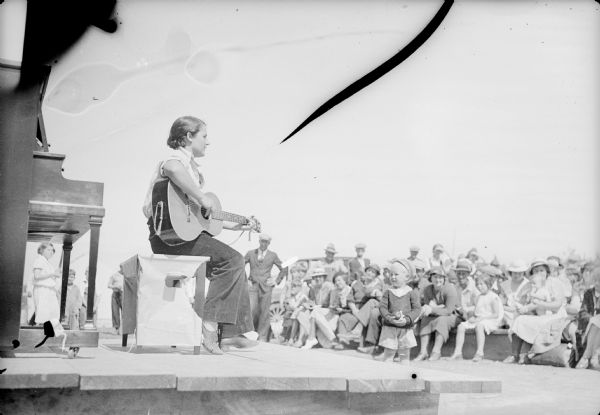 Caption with negative: "Mrs. Walter Fox, one of the colonists, got an encore at the Palmer fourth of July fete by her guitar playing and singing of ballads." Caption on back of print reads: "Home grown entertainment."