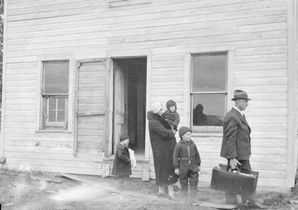 Family, including two children standing, a woman carrying another child, and a man who is carrying suitcases, leaving a house.
