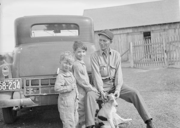 Wesley Worden, Dick and Jackie, and a dog, sitting and standing outdoors at the back of an automobile, which has a Wisconsin license plate. A fence and barn are in the background. Caption with print reads: "Jackie and Dick, talking to their father, Wesley Worden of Three Lakes, Wis., (with dog 'Nerts') after hearing at their farm that they had been selected to be pioneers."