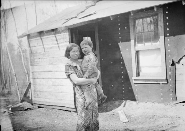 A woman is standing outdoors holding a child in her arms. There is a building in the background. Caption with print reads: "Mrs. Martin Soyk and Jimmy, in front of home they left at Minocqua Wis., to find a better life in Matanuska Colony."