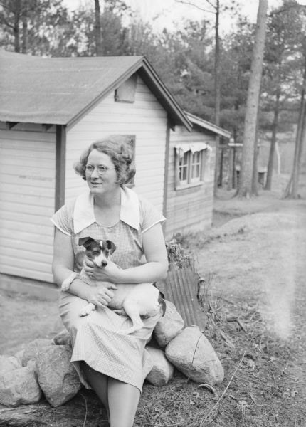 Caption with negative reads: "Mrs. Victor Johnson and Nerts." There are buildings in the background. Caption with print reads: "Mrs. Victor Johnson, from Wisconsin, and her husband choose a house plan of sawed lumber. Later many other colonists choose lumber over logs, as winter approached, because the construction came faster."