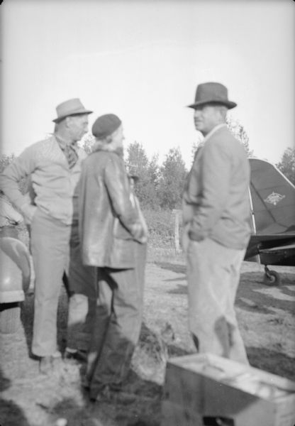 Caption with negative reads: "Rogers talking with Mrs. I.M. Sandvik, colonist from Moose Lake, Minn. In back of Rogers is Stewart Campbell, property custodian on the project." The back end of an airplane is in the background on the right.
