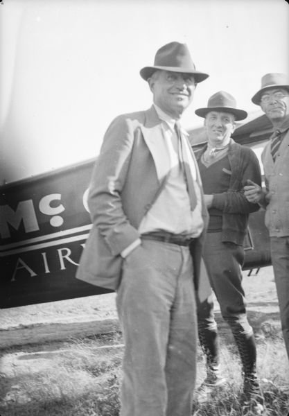 Will Rogers in foreground, with two men standing behind him on the right, and the airplane in the background. Caption with negative: "Rogers alighting from the plane, He is greeted by officials (Col. L.P. Hung and Gene Carr, but I don't know where they are in the picture) and a swarm of transient workers from construction camp."