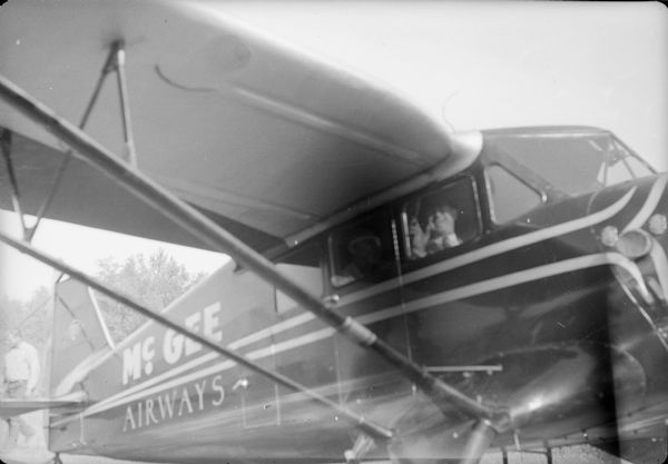Caption with negative: "Immediately after Post and Rogers landed with Pilot Chet McLean of Anchorage, with Post in the front seat with the pilot. Chief Pilot Joe Crosson of the Pacific Alaska Airways also accompanied them." This sign on the side of the plane reads: "McGee Airways." A man is standing in the background behind the airplane on the left.